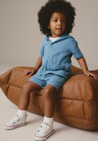 Blå kortærmet skjorte og matchende shorts lavet af tekstureret stof, med hætte og frontlomme. Hvide sneakers med Velcro-stropper.