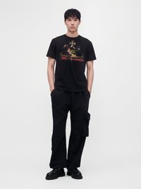 Jeune homme debout portant un t-shirt graphique noir, un pantalon cargo noir ample et des chaussures noires sur un fond blanc uni.