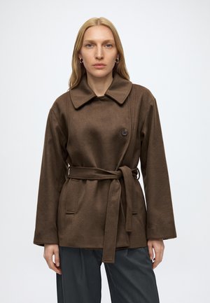 Femme blonde porte un manteau marron ceinturé avec un large col et un pantalon foncé, debout devant un fond clair uni, face à l'avant.