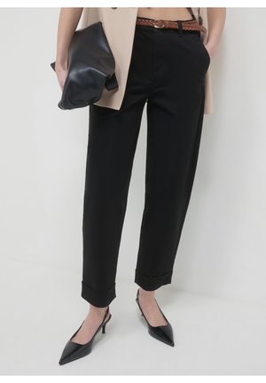 Frau trägt schwarze verkürzte Hose, braunen geflochtenen Gürtel, schwarze spitze Absätze und hält eine weiche schwarze Clutch.