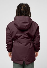 Giacca impermeabile bordeaux con cappuccio, vita elastica e polsini regolabili. Presenta un orlo spaccato per la libertà di movimento. Tessuto liscio.
