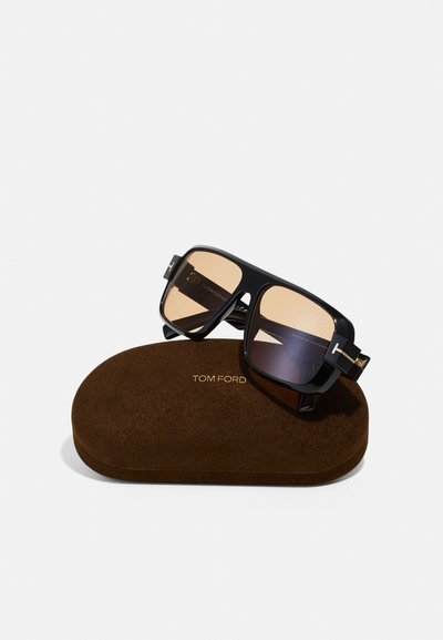 Gafas de sol cuadradas negras con lentes degradados marrones descansando sobre un estuche de ante marrón oscuro. Logo dorado "TOM FORD" en el estuche.