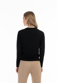 Maglione nero a maglia con scollatura rotonda, polsini aderenti e orlo a coste, abbinato a pantaloni beige. La vista posteriore mette in evidenza una texture liscia.