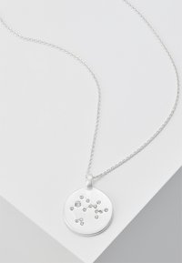 Silberne Halskette mit einem runden Anhänger mit gebürsteter Oberfläche und verstreuten klaren Edelsteinen, die ein Sternenbildmuster bilden, an einer filigranen Kette.