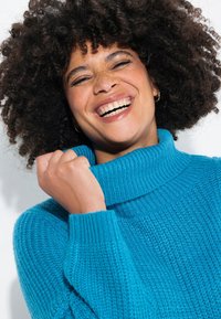 Pull col roulé en tricot turquoise avec un motif texturé. Le modèle est partiellement visible, mettant en avant les poignets et le col du pull.