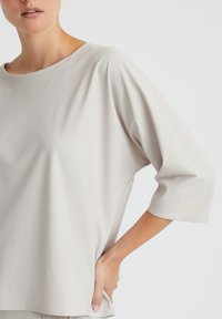 Hellgraues, locker sitzendes Oberteil mit weitem Ausschnitt und Dreiviertelärmeln. Glatter, weicher Stoff mit minimalen Nähten und einer entspannten Silhouette.