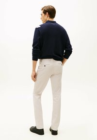 Marine trui, lichtbeige broek, slim fit. Broek heeft twee achterzakken met knoopsluiting. Model draagt zwarte schoenen, gezien van achteren.
