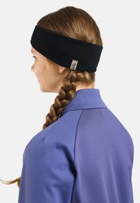 Un bandeau en tissu noir au design plat, avec une petite étiquette. En arrière-plan, une chemise violette. Profil latéral d'une personne avec des cheveux tressés.