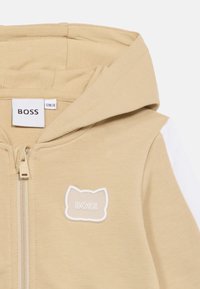 Μπεζ φούτερ με φερμουάρ, άσπρη ρίγα στο μανίκι, μπαλώμα σε σχήμα γάτας με την ετικέτα «BOSS» και ταμπέλα επωνυμίας που δείχνει μέγεθος 12Μ-74.