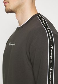 Uomo che indossa un felpa grigio scuro con il logo bianco "Champion" sul petto e una striscia nera sulla spalla con il marchio "Champion" ripetuto.