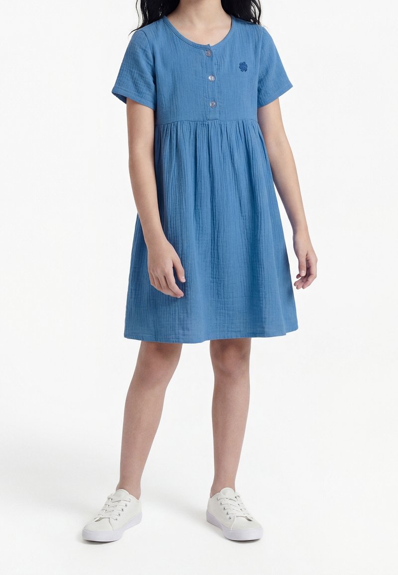Fille portant une robe bleue mi-longue à manches courtes avec des boutons sur la poitrine, associée à des baskets blanches, debout sur un fond blanc.