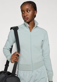 Světlá zelená bunda na zip z měkkého materiálu, s vysokým límcem, žebrovaným lemem a odpovídajícími světlými kalhotami. Drží černou tašku.