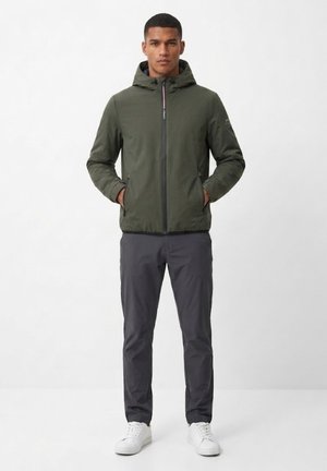 Hombre de pie con las manos en los bolsillos, vestido con una chaqueta con capucha verde oliva, pantalones gris oscuro y zapatillas blancas sobre un fondo liso.