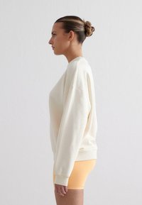 Felpa oversize color crema con spalle abbassate e polsini a coste, abbinata a pantaloni corti arancioni chiari, mostrata di lato.