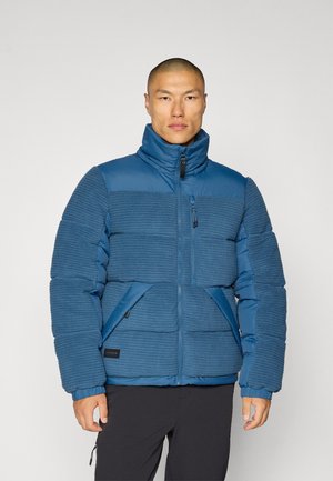 Blue padded jacket with horizontal textured stripes, high collar, full zipper, and side pockets. Features a matte nylon finish and branding patch.