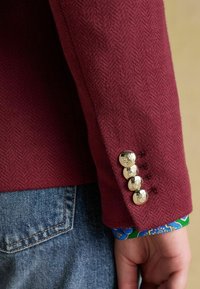 Bordeaux-tooni herringbone musteriga bleiseri varrukas on kuldne nupp ja sinine mustriline mansett nähtaval. Bleiseril on risttõmme sobivus ja tekstureeritud kangas.