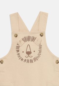 Monos de color beige hechos de algodón con un gráfico de la cara de un león y detalles de orejas, cuentan con botones a presión de latón y bolsillos frontales.