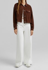 Kratká hnědá manšestrová bunda se dvěma předními kapsami, v kombinaci s bílými širokými kalhotami s vysokým pasem a bílými teniskami.