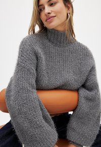 Grauer, flauschiger Rollkragenpullover mit lockeren Ärmeln, der ein strukturiertes Strickdesign aufweist. Weicher Stoff und lockere Passform. Tiefer Ausschnitt und gerippte Details.