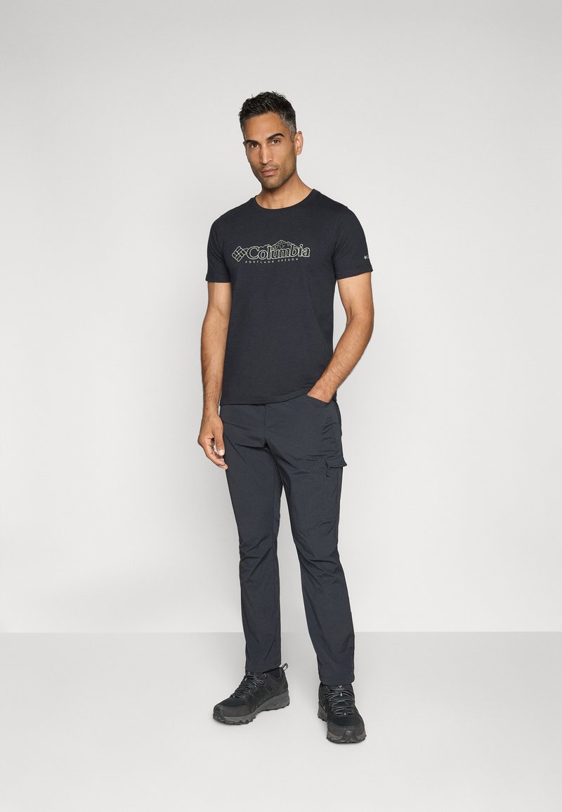 Schwarzes T-Shirt mit grünem Logo, kombiniert mit dunklen Cargo-Hosen und robusten schwarzen Schuhen. Das Modell steht vor einem schlichten hellen Hintergrund.