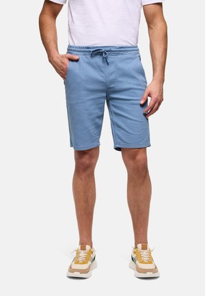 Hombre con pantalones cortos azul claro con cordón, camiseta blanca y zapatillas beige con detalles en amarillo y negro, de pie con una mano en el bolsillo.