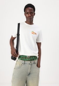 Λευκό βαμβακερό μπλουζάκι με γραφικό ροδάκινο στο στήθος, συνδυασμένο με ανοιχτό μπλε τζην σορτς και πράσινα καρώ μποξεράκια. Μαύρη τσάντα ώμου.