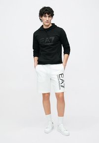 Svart hettegenser med EA7-logo, hvite shorts med logodetalj, hvite joggesko og hvite sokker. Enkel design, bomullsmateriale og avslappet passform.