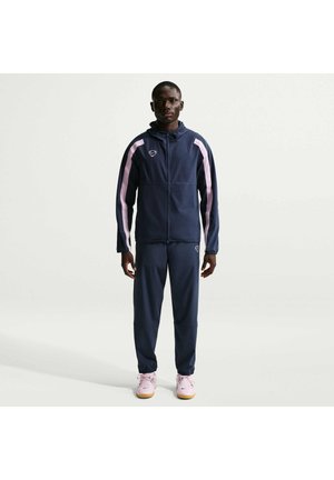 Homme debout, faisant face à l'avant, portant un survêtement bleu marine avec des rayures roses sur les manches et les jambes, et des baskets rose clair, fond neutre.