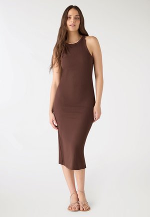 Giovane donna con lunghi capelli castani che indossa un vestito midi marrone senza maniche e sandali piatti beige con cinturini, in piedi davanti a uno sfondo bianco.