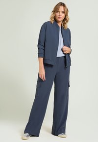 Marineblauwe bomberjack met geribbelde boorden, gecombineerd met bijpassende wide-leg broek met cargozakken. Gladde stof met een ontspannen pasvorm.