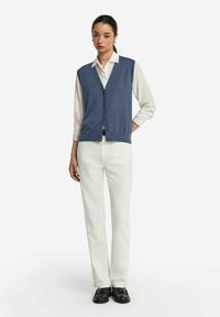 Marinblå stickad väst med v-ringning, knäppning framtill, bärs över en vit skjorta med krage, kombinerad med vita raka byxor och svarta skor.