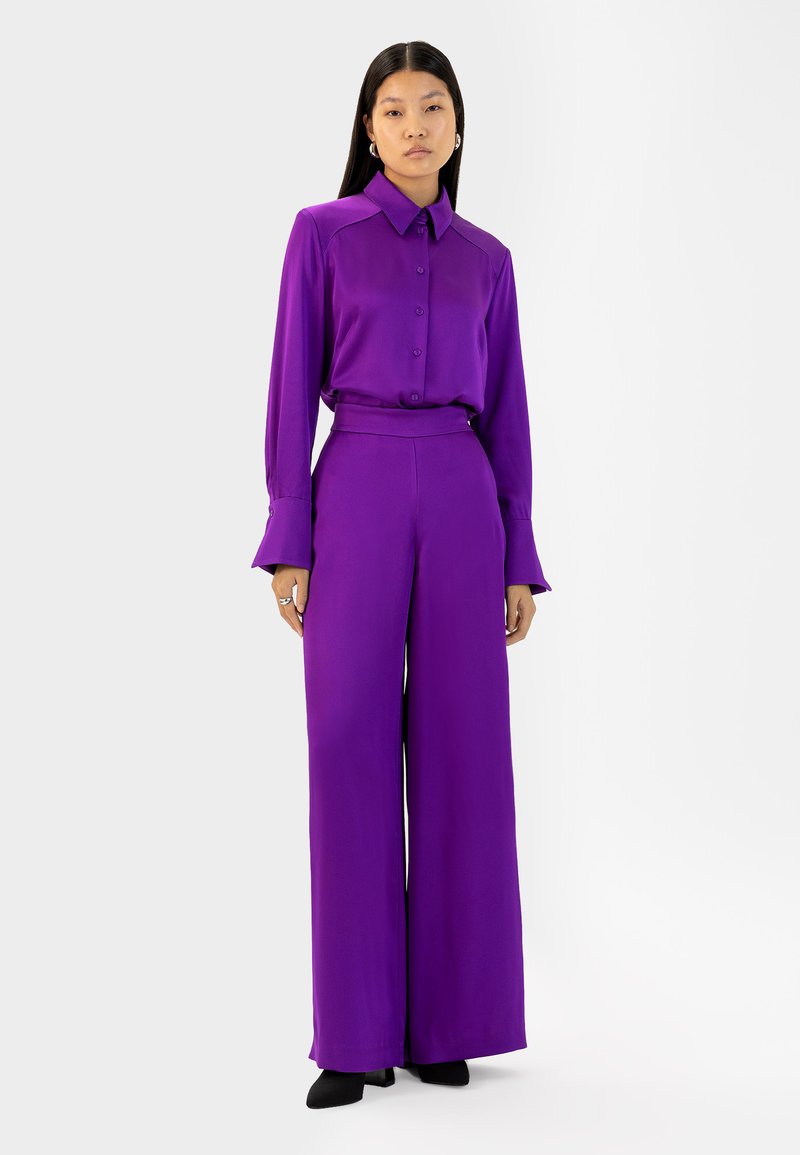 Paarse blouse met knoopsluiting aan de voorkant en brede manchetten, gecombineerd met hoog-waist, brede paarse broek. Glad stof en schone lijnen overal.