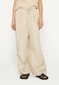 Pantaloni beige a gamba larga con vita elasticizzata e coulisse, caratterizzati da pieghe e una texture liscia, indossati con sandali intrecciati neri.