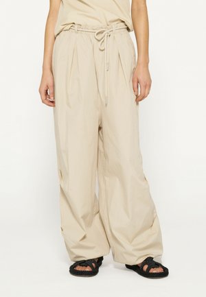 Pantaloni beige a gamba larga con vita elasticizzata e coulisse, caratterizzati da pieghe e una texture liscia, indossati con sandali intrecciati neri.