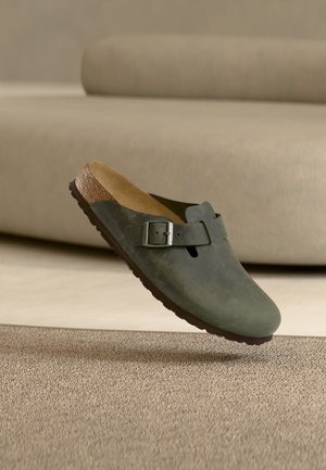 Zueco verde sin cordones con hebilla ajustable, plantilla de corcho y suela de goma sobre una alfombra beige con un sofá beige de fondo.