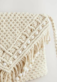 Beige gehäkelte Handtasche mit strukturiertem Oberflächendesign, verziert mit weißen Perlenakzenten und Fransen am unteren Rand. Verstellbarer Tragegurt inklusive.