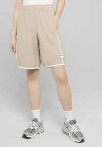 Beige knit shorts with an elastic waistband, side pockets, white piping, and a logo patch on the hem. Worn with gray sneakers and white socks.