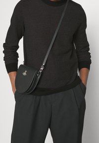 Maglione nero a maglia con texture a coste e pantaloni neri plissettati. Piccola borsa a tracolla nera con forma curvata e accentuata da un logo metallico.