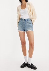 Cárdigan de punto en beige claro sobre una camiseta blanca de tirantes, combinado con pantalones cortos de mezclilla desgastados, mocasines negros y calcetines blancos hasta el tobillo.