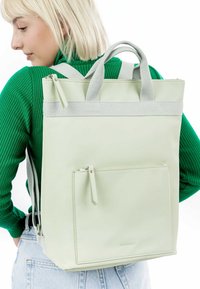 Lichtgroene tote rugzak van glad materiaal, met grijze banden, een bovenhandvat en een voorzak met rits voor gemakkelijke toegang.