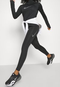 Černý sportovní outfit skládající se z dlouhého rukávu s vysokým límcem a legín, oba s reflexními logy Nike, doplněný o černé tenisky.