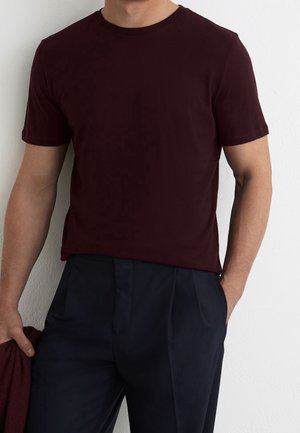 Mężczyzna noszący gładki, ciemny bordowy T-shirt z krótkim rękawem i ciemne granatowe spodnie, trzymający w lewej ręce pasujący bordowy materiał, na tle białej ściany.