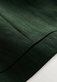 Mørkegrønt stof med en struktureret overflade, der har en diagonal søm og bløde ribbede kanter. Materialet fremstår holdbart og fleksibelt.