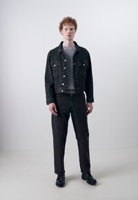Sort denimjakke med sølvknapper lagdelt over en grå ribbet sweater, parret med matchende sorte denimbukser og sorte sko.