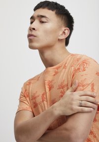Kortermet oransje T-skjorte med teksturert overflate, prydet med et rødt plantebladmønster, rund hals og avslappet passform.