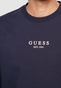 Námořnická modrá mikina vyrobená z bavlny, s žebrovaným kulatým výstřihem a bílým vyšitým logem "GUESS EST.1981" na hrudi.