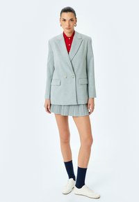 Lichtgrijze dubbele blazer met notch-kraag, gecombineerd met een geplooide rok. Rode blouse met kraag, witte sneakers en marineblauwe kniekousen.