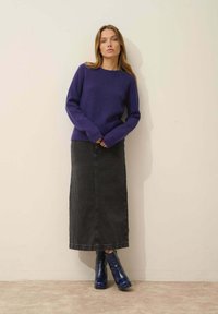 Lila gerippter Strickpullover, schwarze Denim-Maximum-Rock und marineblaue Stiefeletten mit glänzender Textur. Stehend vor einer hellen Wand.