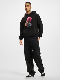 Sort hættetrøje med et grafisk design, der viser et farverigt hoved. Materialet ser blødt ud. Sammen med sorte løse jeans og sorte sneakers.