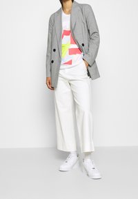 Blazer de cuadros gris de doble botonadura sobre una camiseta gráfica blanca con formas geométricas en rosa, rojo y amarillo. Combinado con pantalones anchos blancos y zapatillas.